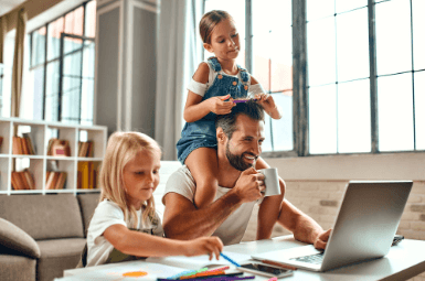 A father works on a laptop at home while his two young daughters play beside him, one sitting on his shoulders and the other drawing at the table.