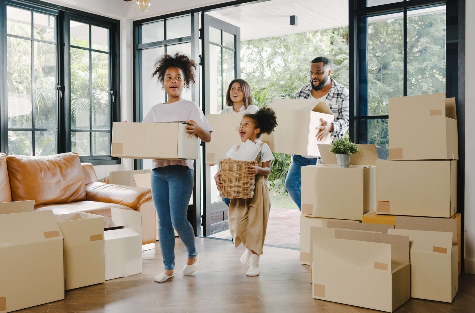 A family carries moving boxes into their new home, smiling as they enter a bright living room filled with packed boxes and natural light from large windows.