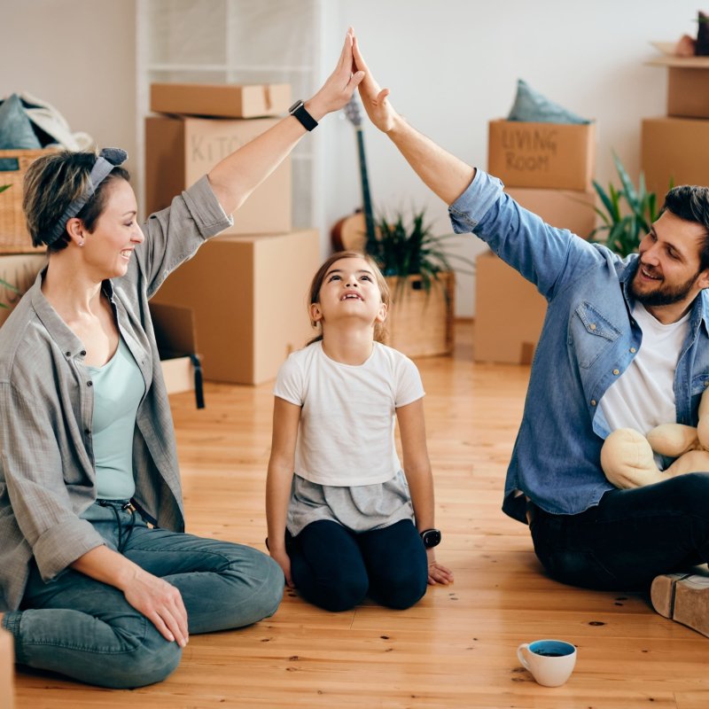 A happy family sits on the floor surrounded by moving boxes, smiling and giving a high-five to celebrate moving into their new home.
